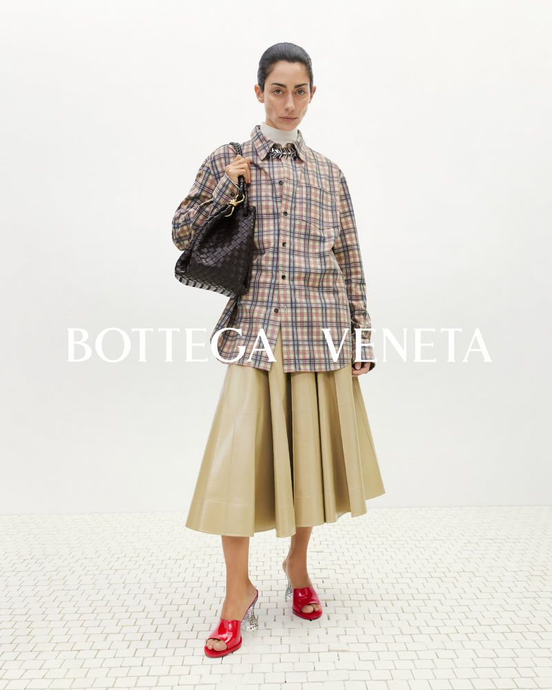 Fashionable woman in plaid shirt and tan skirt poses indoors with a black woven handbag and red mules, logo in foreground.