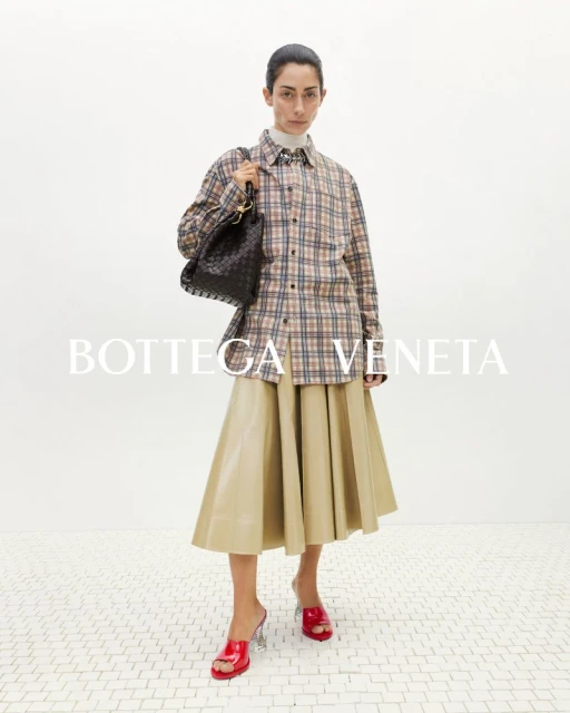 Fashionable woman in plaid shirt and tan skirt poses indoors with a black woven handbag and red mules, logo in foreground.
