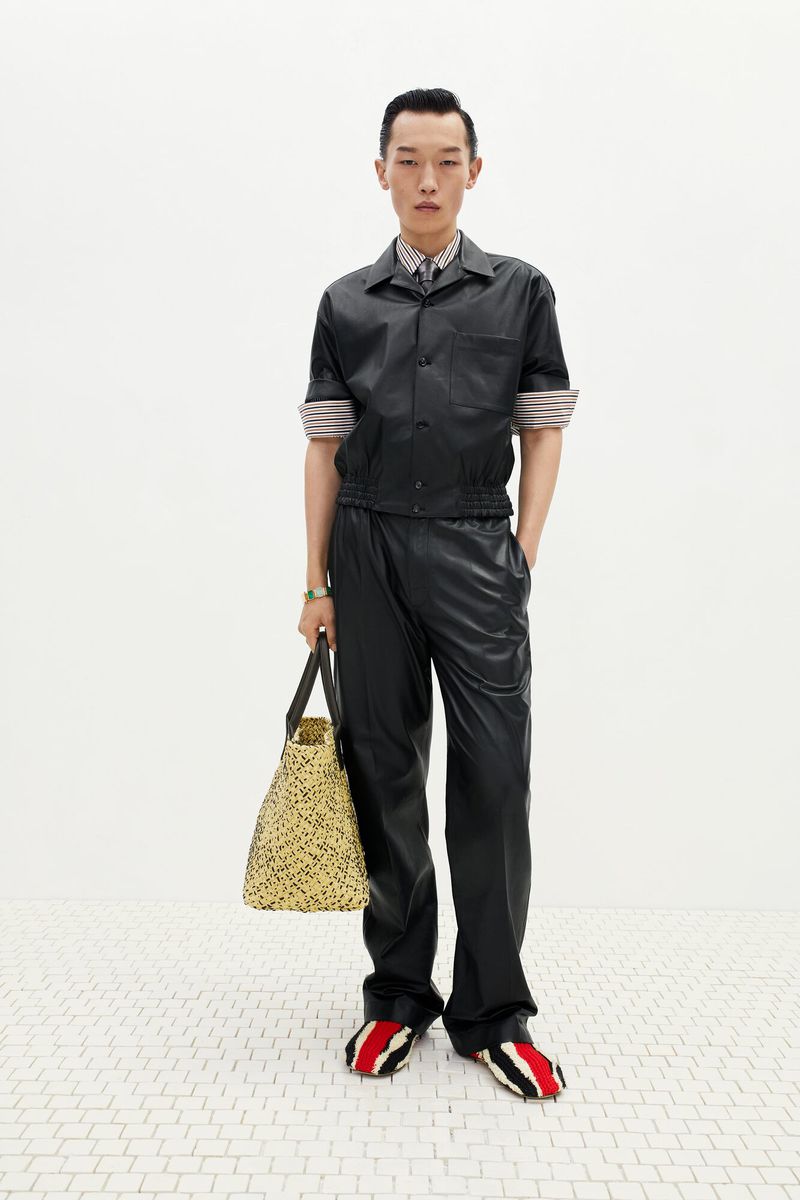 Asian man poses in a black leather-like shirt, black pants, a neutral bag, and red-striped shoes against a white backdrop and tiled floor.