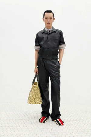 Asian man poses in a black leather-like shirt, black pants, a neutral bag, and red-striped shoes against a white backdrop and tiled floor.