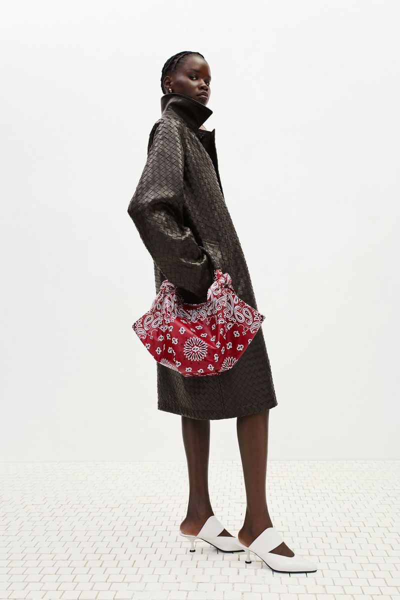 A fashion shot features a Black female model in a textured coat, accessorized with a patterned handbag and white mule shoes, standing indoors.