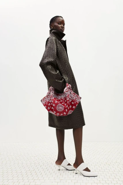 A fashion shot features a Black female model in a textured coat, accessorized with a patterned handbag and white mule shoes, standing indoors.