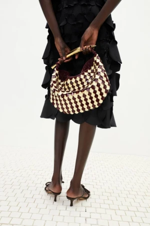 Fashionable woman showcasing a woven handbag and bow-detail heels, against a white backdrop. Dark-skinned model wearing a black dress.
