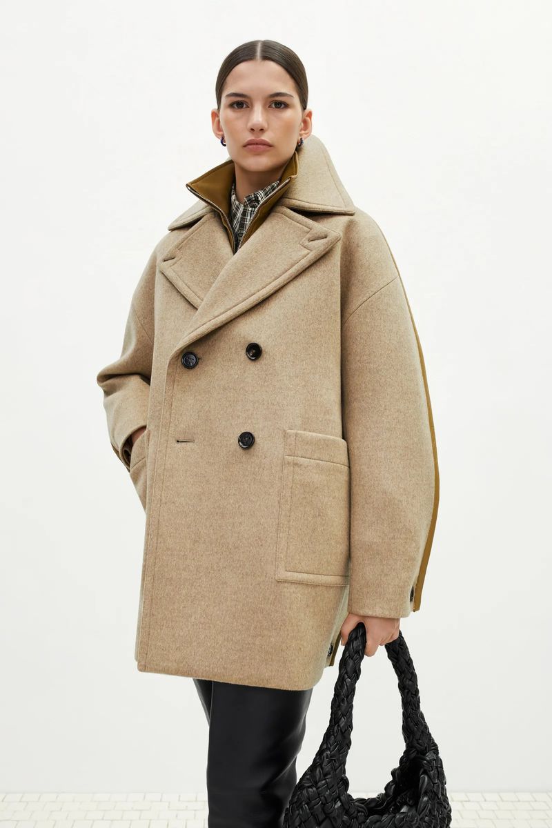 Model wearing an oversized beige double-breasted coat and holding a black woven leather bag against a white studio background.