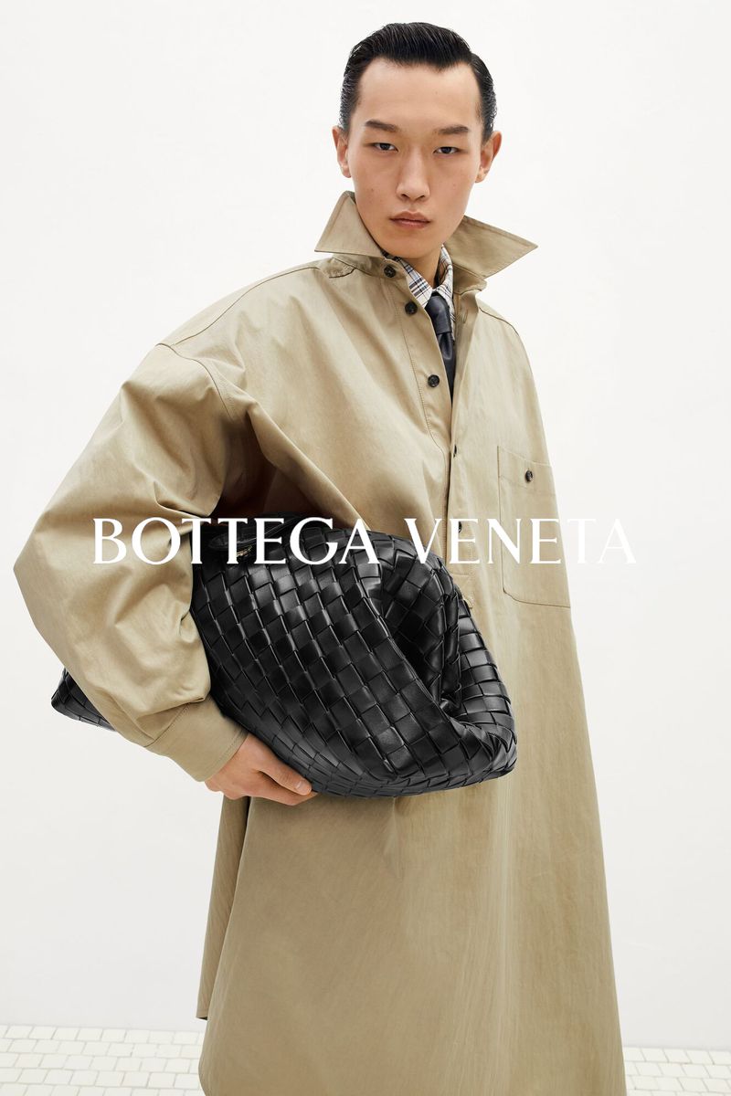 An East Asian male model wearing a light beige coat holds a large black woven leather bag against a white backdrop.