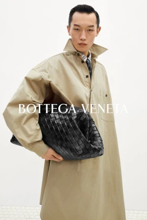 An East Asian male model wearing a light beige coat holds a large black woven leather bag against a white backdrop.