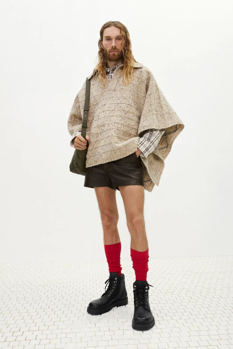 Full-length studio shot of a man in a poncho, shorts, boots, and red socks, standing on a white tiled floor against a white background.