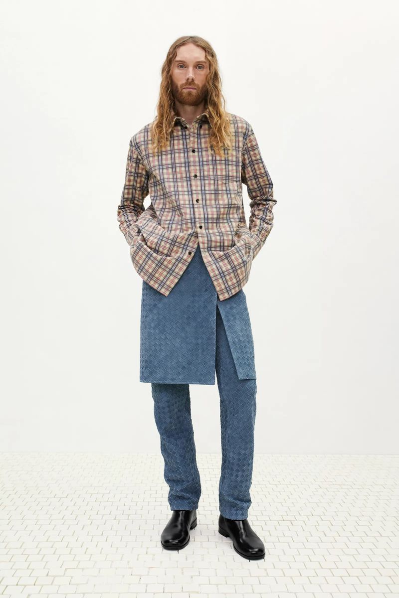 Male fashion model posing in a full shot in a plaid shirt, textured jeans and an asymmetrical wrap in a bright, neutral studio.