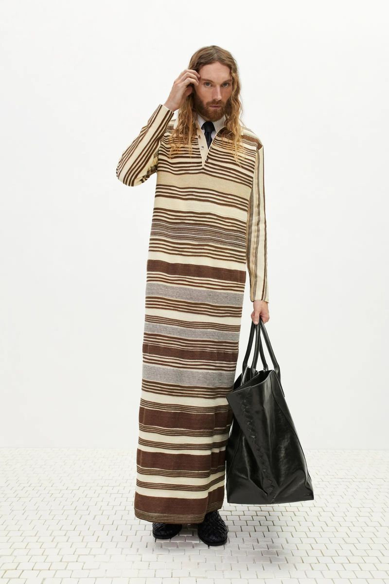 Male model in striped maxi dress holding black leather tote bag in a bright studio setting, fashion editorial style, full body shot.