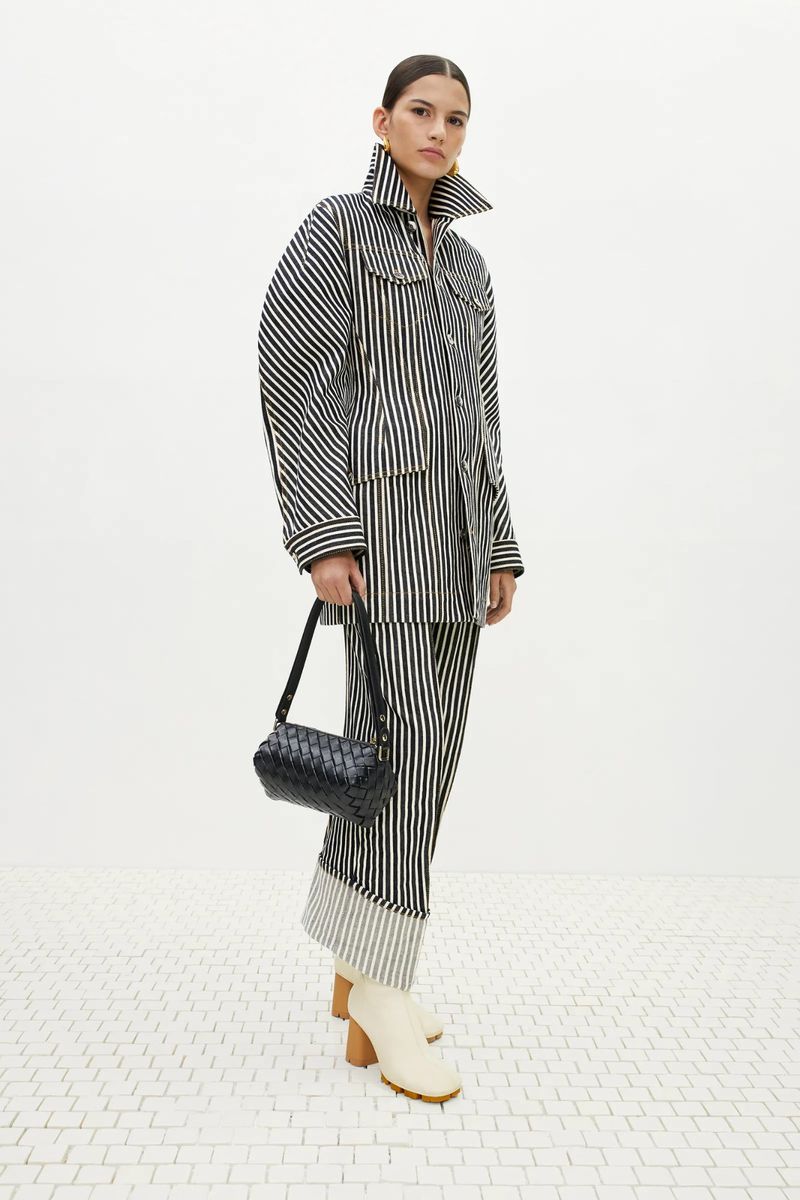 Adult woman in a black and white striped jacket and pants, holding a black purse, posing in a light, neutral environment.