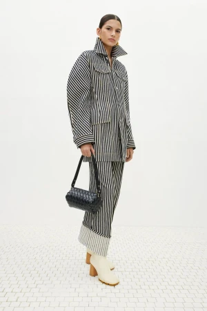 Adult woman in a black and white striped jacket and pants, holding a black purse, posing in a light, neutral environment.