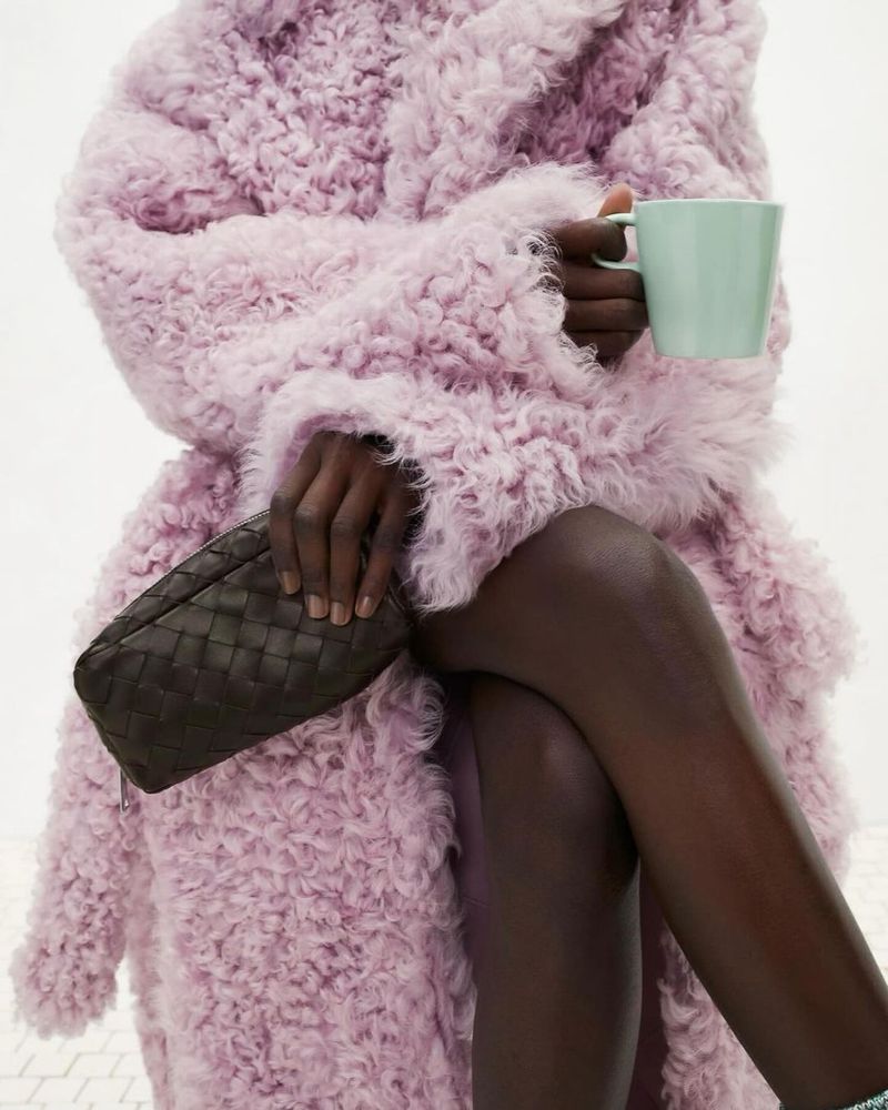 An adult woman is seated and wearing a fluffy lilac coat, while holding a clutch and a mug in an indoor setting.