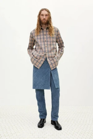 Man with long wavy hair wearing a plaid shirt, layered blue skirt, and trousers against a white background.