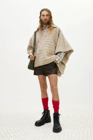 Model wearing a beige speckled knit poncho, dark leather shorts, red socks, and black boots for Bottega Veneta Pre-Fall 2024.