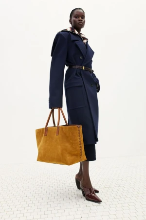 Model wearing a navy belted overcoat and burgundy heels holding a large mustard yellow suede tote against a white backdrop.