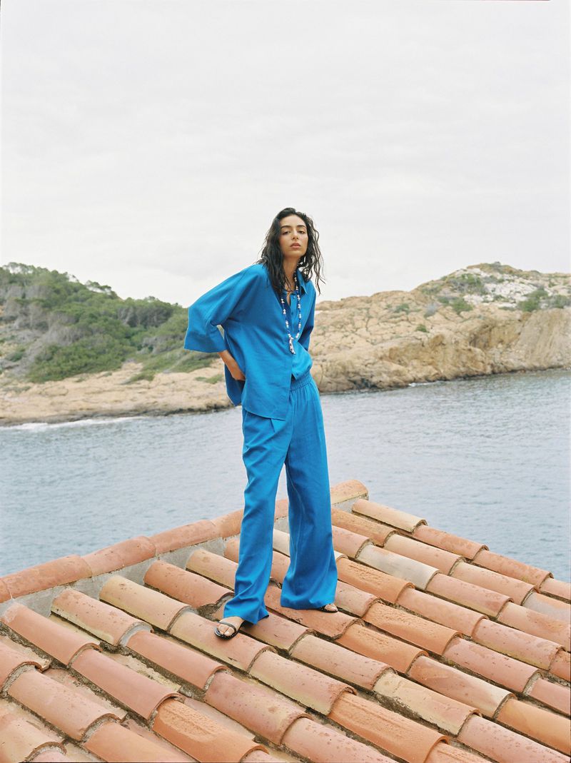 Woman in a turquoise blouse and wide-leg pants poses on a terracotta tile rooftop overlooking the ocean and a rocky, green coastline.