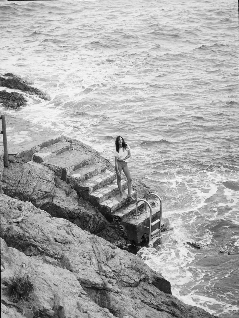 Black and white photograph of a woman standing on concrete stairs with a metal handrail, leading into the ocean with rocky cliffs.