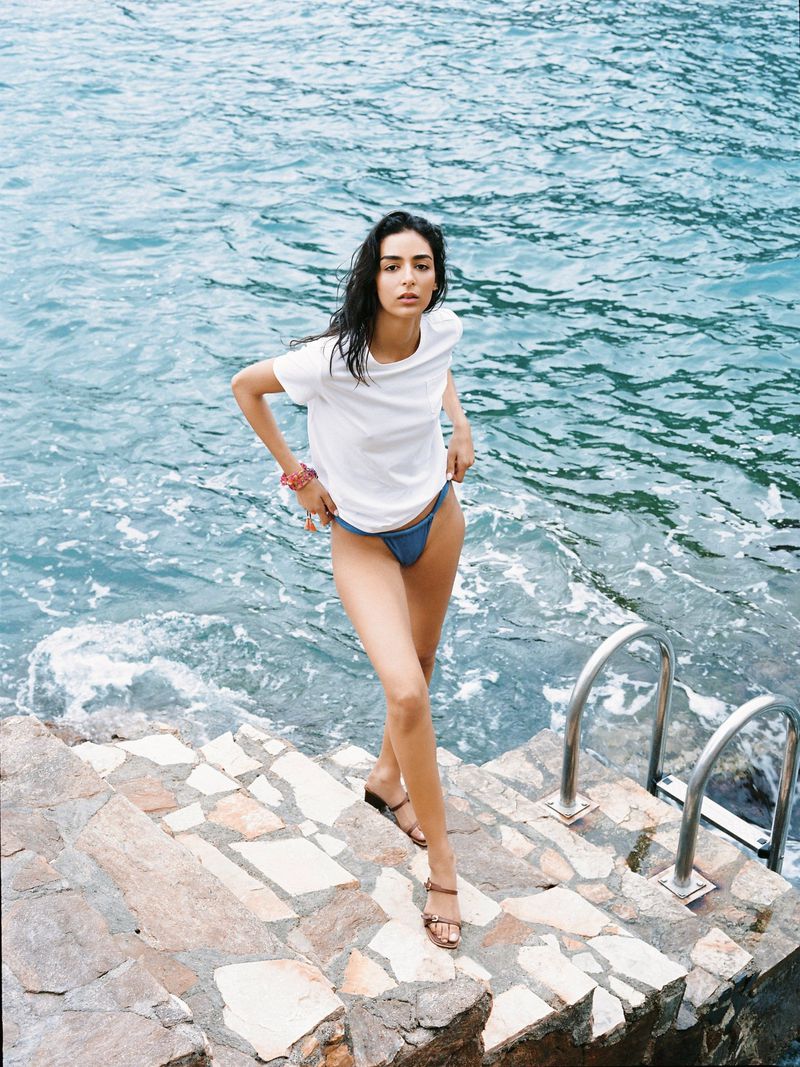 An attractive woman walks down stone steps near the ocean. She wears a t-shirt and swimsuit bottoms in a daylight, outdoor fashion shot.