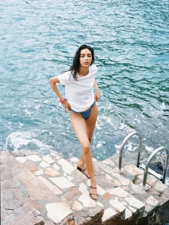 An attractive woman walks down stone steps near the ocean. She wears a t-shirt and swimsuit bottoms in a daylight, outdoor fashion shot.