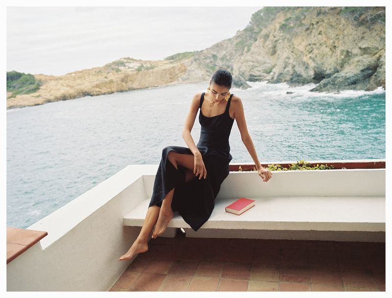 A brunette woman in a black dress sits on a white bench with a red book beside her, overlooking the ocean with rocky cliffs.