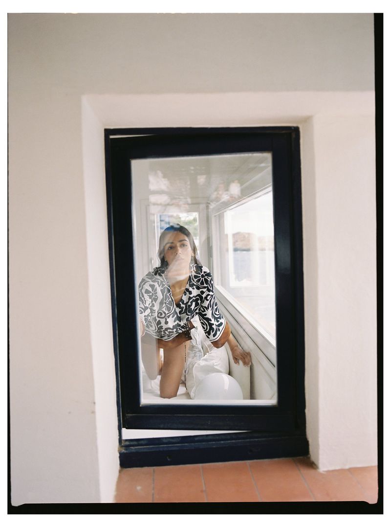 Indoor portrait of an adult woman in a patterned top, kneeling in a window alcove with a white bag, captured in soft, natural light.
