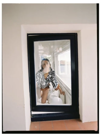 Indoor portrait of an adult woman in a patterned top, kneeling in a window alcove with a white bag, captured in soft, natural light.