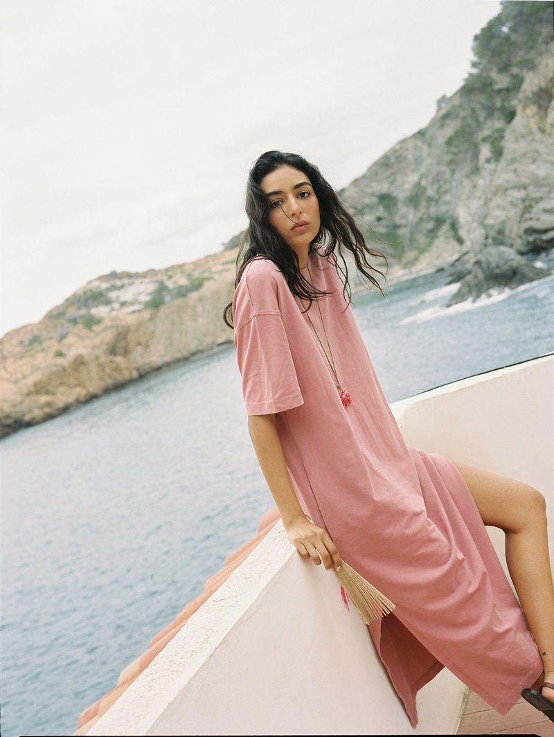 A brunette woman in a pink dress sits on a wall overlooking the ocean, with a fan in her hand, posing outdoors on a sunny day.