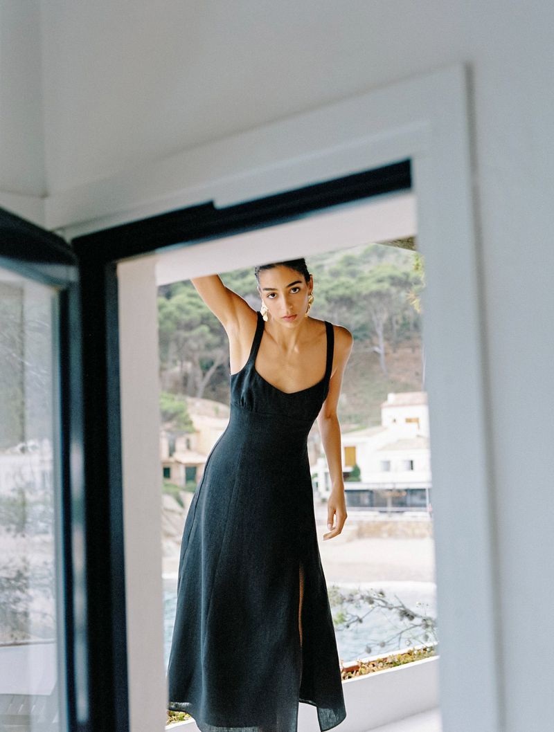 A young woman in a navy midi sundress stands in an open white doorway, looking at the camera with a beach and Mediterranean village view.