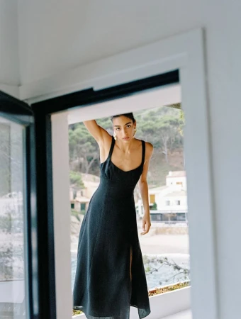 A young woman in a navy midi sundress stands in an open white doorway, looking at the camera with a beach and Mediterranean village view.