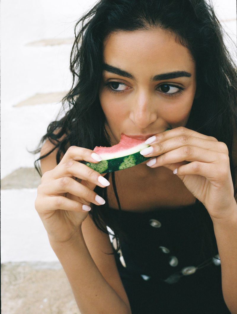 Woman with dark hair eats watermelon. Close-up of dark-haired person in summer clothing enjoying fresh fruit, set against light stairs.