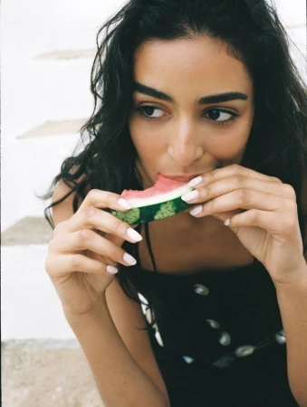 Woman with dark hair eats watermelon. Close-up of dark-haired person in summer clothing enjoying fresh fruit, set against light stairs.