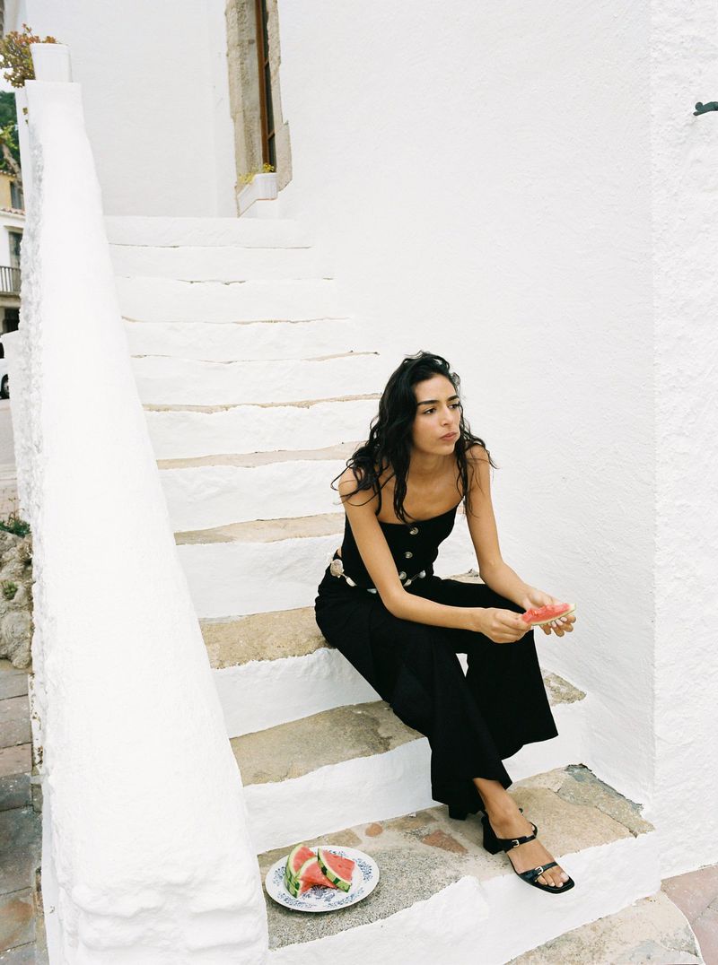 Brunette woman in a black outfit sits on white steps, holding a slice of watermelon with plate of watermelon slices nearby.
