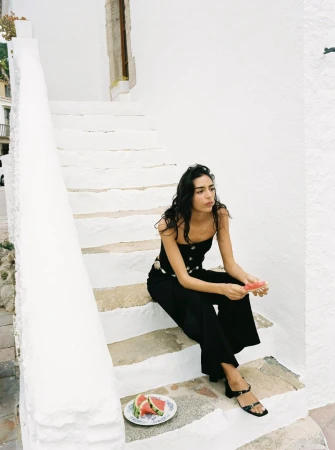Brunette woman in a black outfit sits on white steps, holding a slice of watermelon with plate of watermelon slices nearby.
