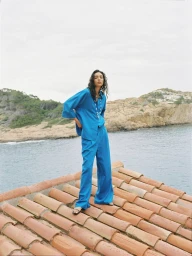 Model Nora Attal wearing a blue shirt and trousers standing on a terracotta tiled roof overlooking water and hills.