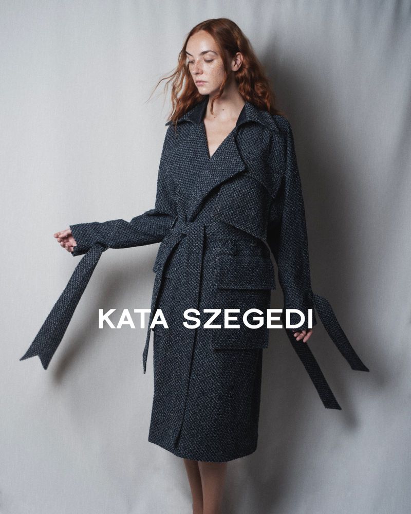 Model wearing a dark textured belted coat against a neutral backdrop for Kata Szegedi SS 25 lookbook.