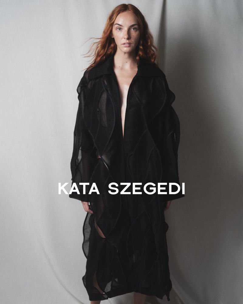 Fashionable red-haired woman wears a long, sheer black jacket in a studio setting, showcasing contemporary style and neutral tones.