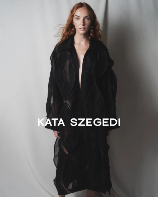 Fashionable red-haired woman wears a long, sheer black jacket in a studio setting, showcasing contemporary style and neutral tones.