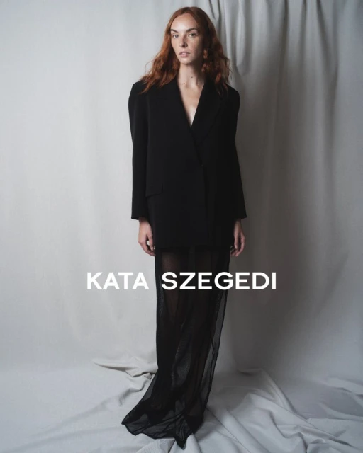 Fashion shot of a red-haired woman posing in a black jacket and black sheer skirt in front of a grey curtain backdrop in studio.