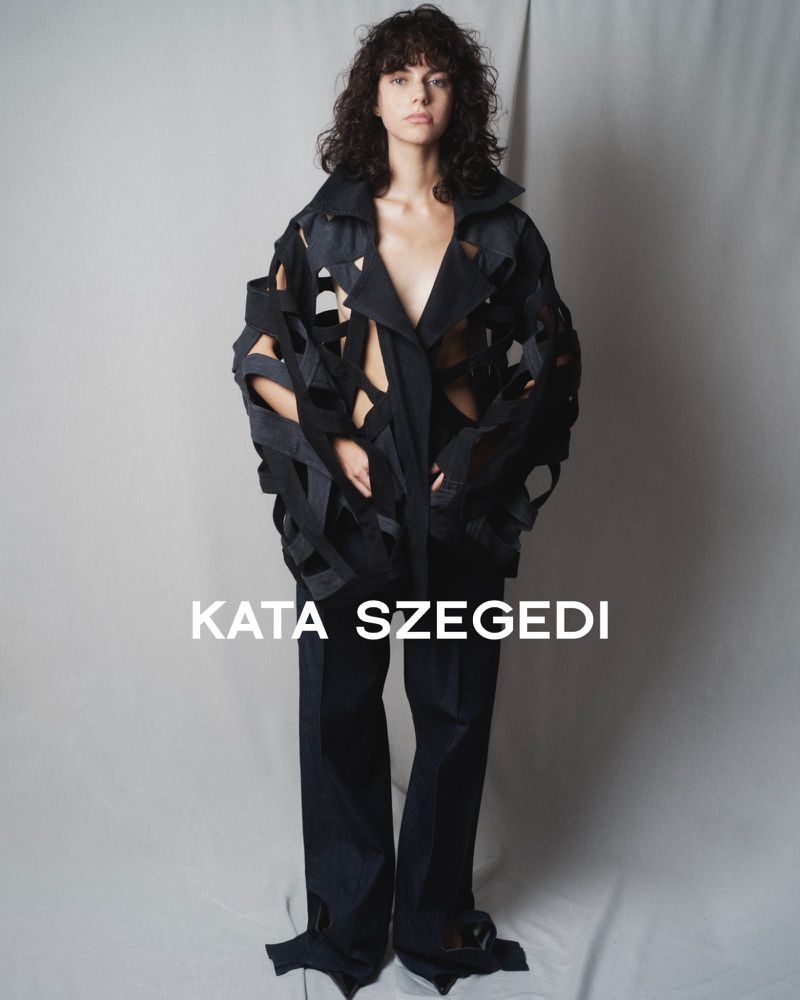 Fashion shot: woman in cut-out jacket and wide-leg trousers poses in a studio setting.  Designer Kata Szegedi’s brand name is visible.