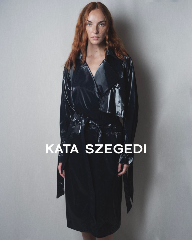 An adult female with red hair poses indoors wearing a shiny black trench coat, standing against a neutral backdrop with 'Kata Szegedi' text.