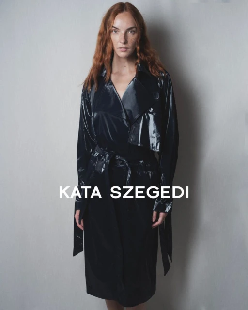 An adult female with red hair poses indoors wearing a shiny black trench coat, standing against a neutral backdrop with 'Kata Szegedi' text.