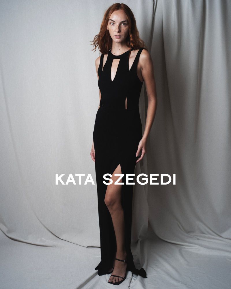 Woman poses in a black Kata Szegedi gown featuring a thigh-high split, cutout details at the bodice, and thin strapped black heels.