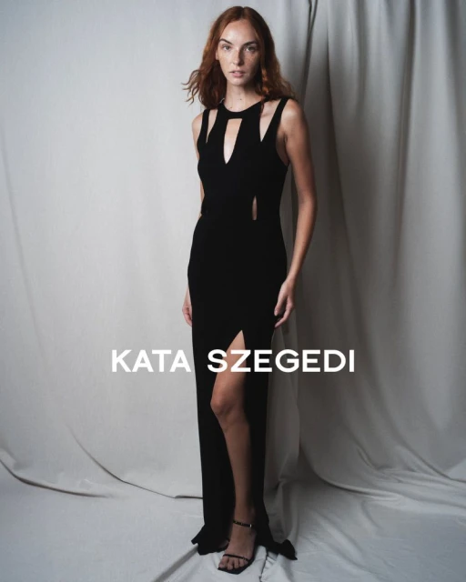 Woman poses in a black Kata Szegedi gown featuring a thigh-high split, cutout details at the bodice, and thin strapped black heels.