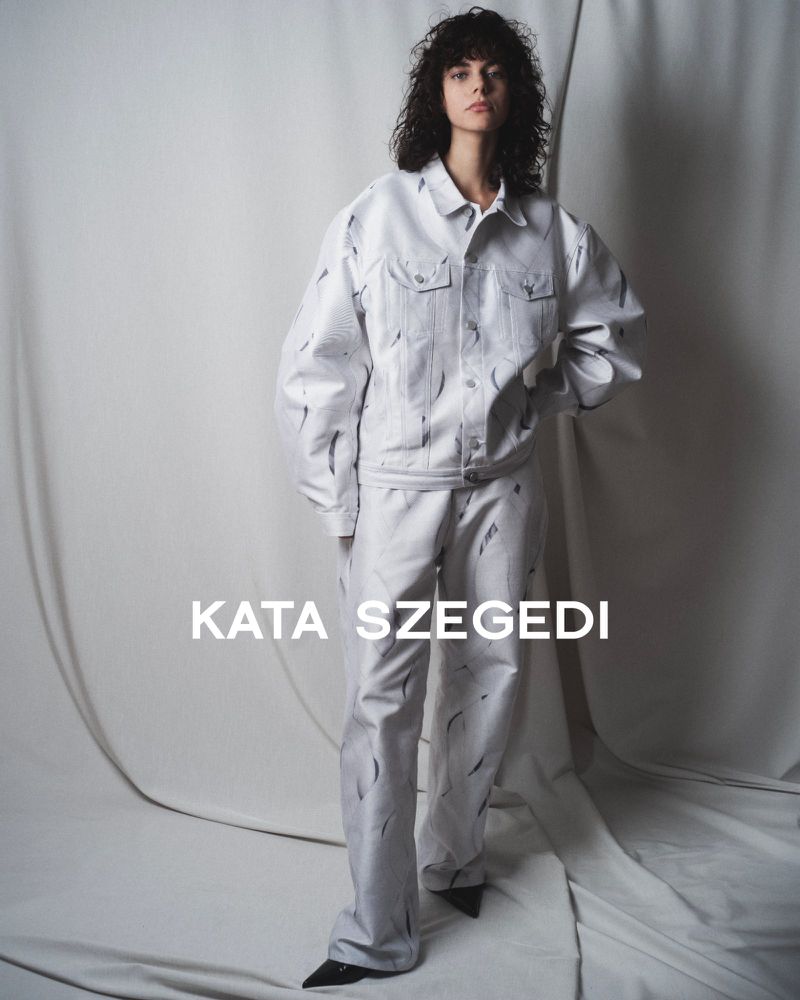 A model wears a matching white denim jacket and trousers with black markings against a neutral studio backdrop for a fashion editorial.
