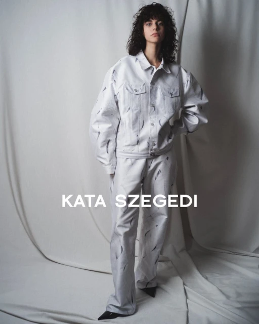 A model wears a matching white denim jacket and trousers with black markings against a neutral studio backdrop for a fashion editorial.