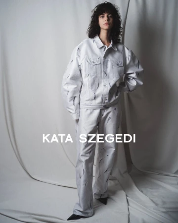 A model wears a matching white denim jacket and trousers with black markings against a neutral studio backdrop for a fashion editorial.