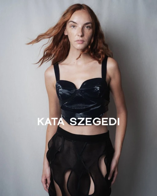 Fashion portrait of a red-haired woman wearing a shiny black top and sheer black skirt against a neutral backdrop in a studio setting.