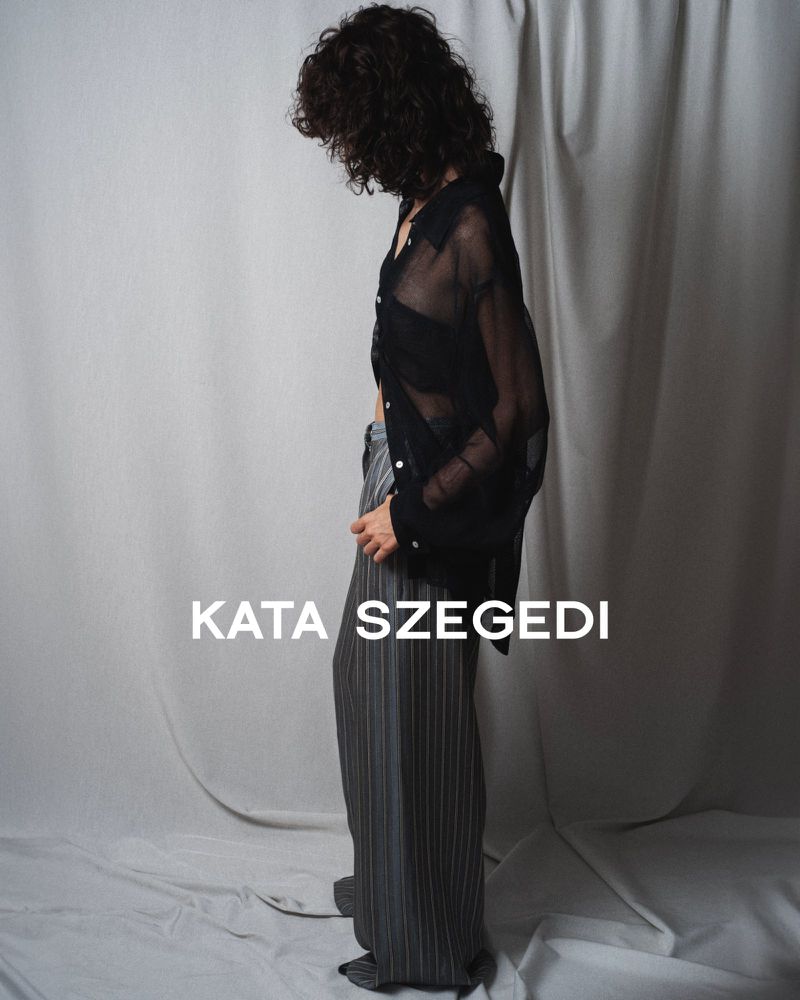 Model with dark curly hair wearing a sheer black button-down shirt and grey pinstripe trousers against a draped grey fabric backdrop.
