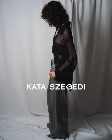 Model with dark curly hair wearing a sheer black button-down shirt and grey pinstripe trousers against a draped grey fabric backdrop.