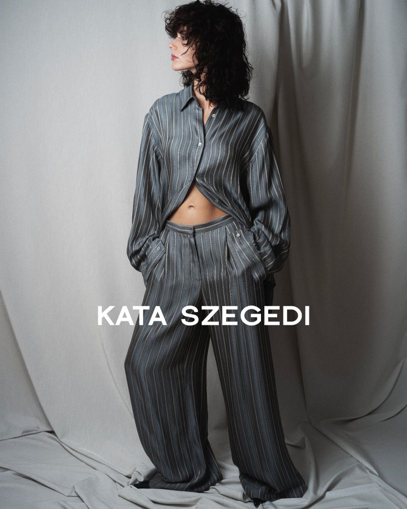 A woman with curly dark hair poses in a grey striped blouse and pants set in a studio with neutral-toned draped backdrop and soft lighting.